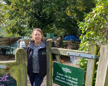 Project Coordinator Frankie Kennet, stands at the gate of a compost demonstration site, next to a sign that reads "come on in and see our compost demonstration site"
