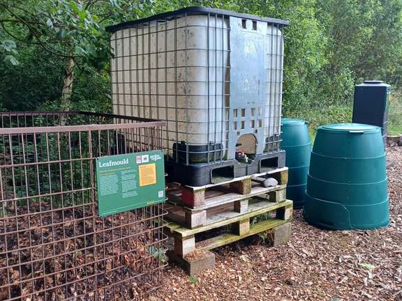 A metal cage with an open top that has piles of leaves next to it. Next to the cage are 3 wooden pallets stacked on top of each other and a Green Johanna compost bin