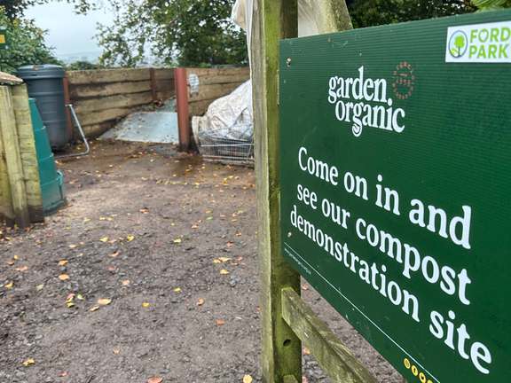 A gate, opened and leading to a compost area. The gate has a dark green sign on, the sign reads "come on in and see our compost demonstration site"